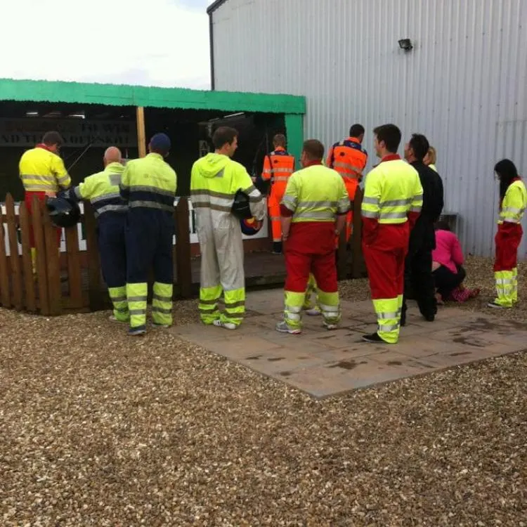 Group of workers and visitors in high-visibility clothing gathered outside a green-roofed facility at a karting venue.