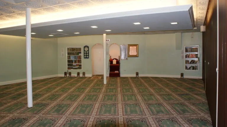 Empty prayer hall with patterned carpet, support pillars, bookshelves and decorative wall features.