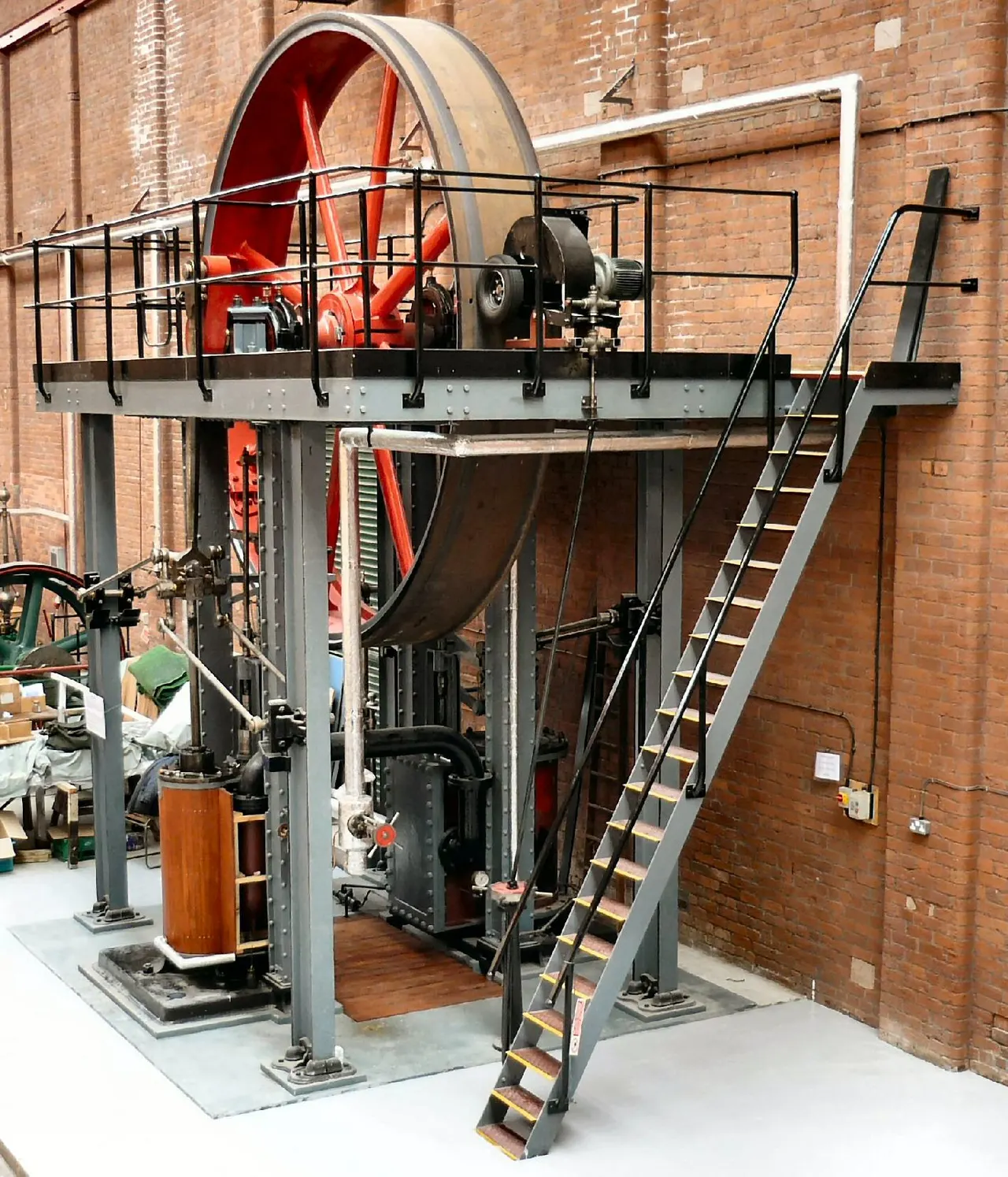 Historic steam engine machinery displayed indoors against a brick building, with red wheel and metal framework structure.