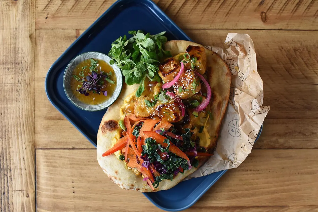 Asian-style bao bun filled with glazed meat, pickled vegetables and fresh herbs, served with dipping sauce on a blue tray.