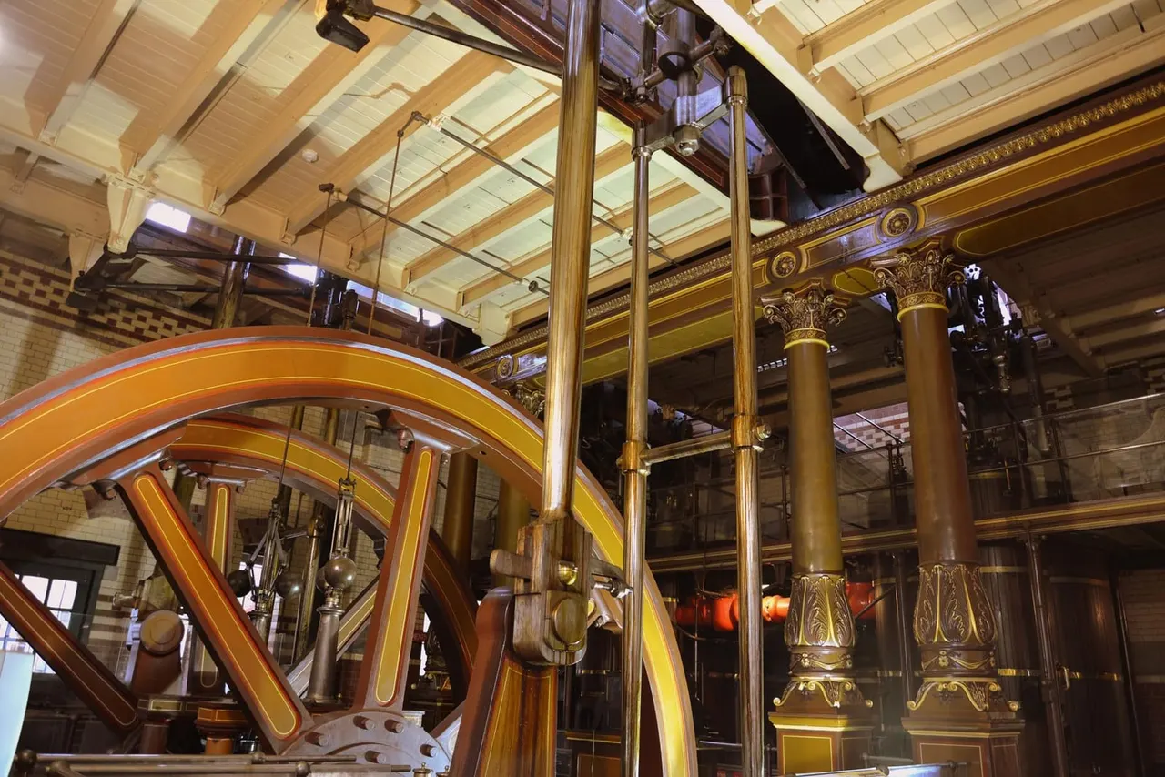Interior of a grand Victorian building with ornate gilt columns, decorative moulding, and a large illuminated wheel mechanism displayed behind glass.
