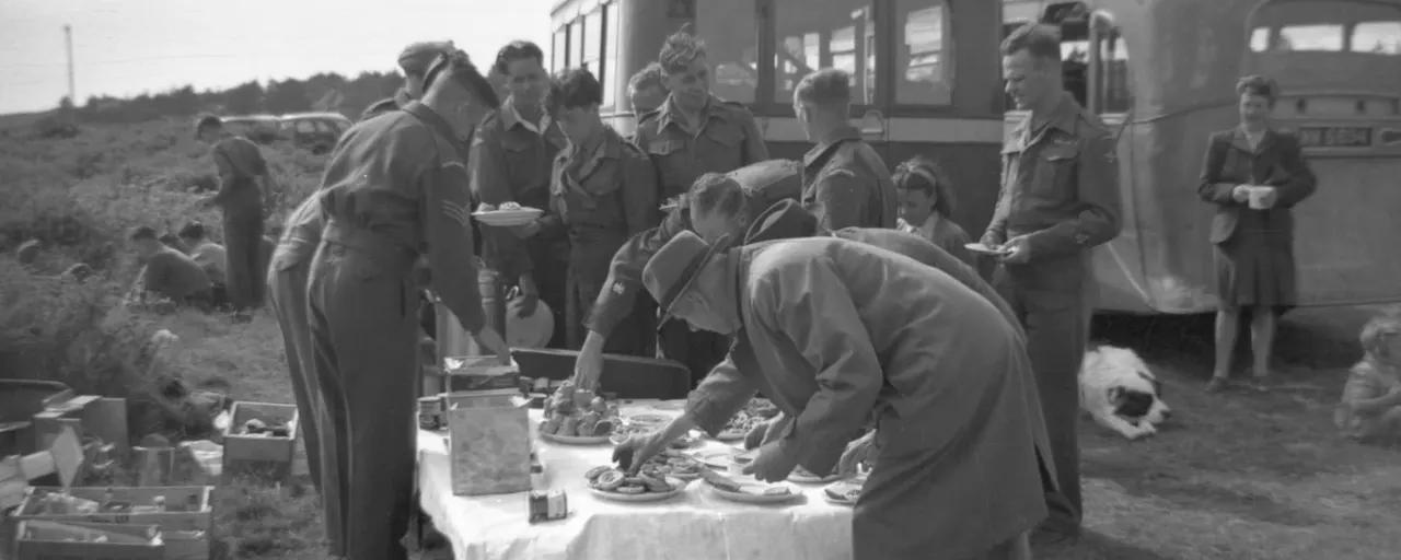 Black and white photograph of soldiers serving food outdoors from a table to a gathered group, with a military vehicle in the background.