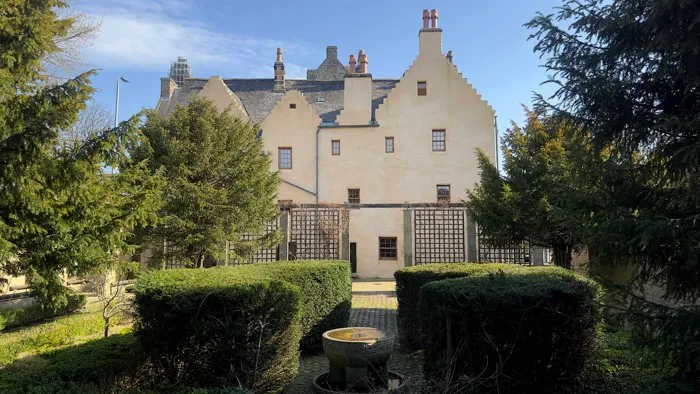 Historic cream-coloured mansion with stepped gable roof viewed from formal garden with hedges and central circular feature.