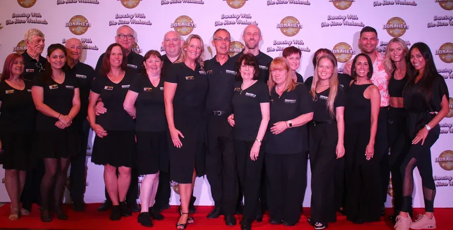 A large group of staff members in black uniforms posing together on a red carpet at a branded event backdrop.
