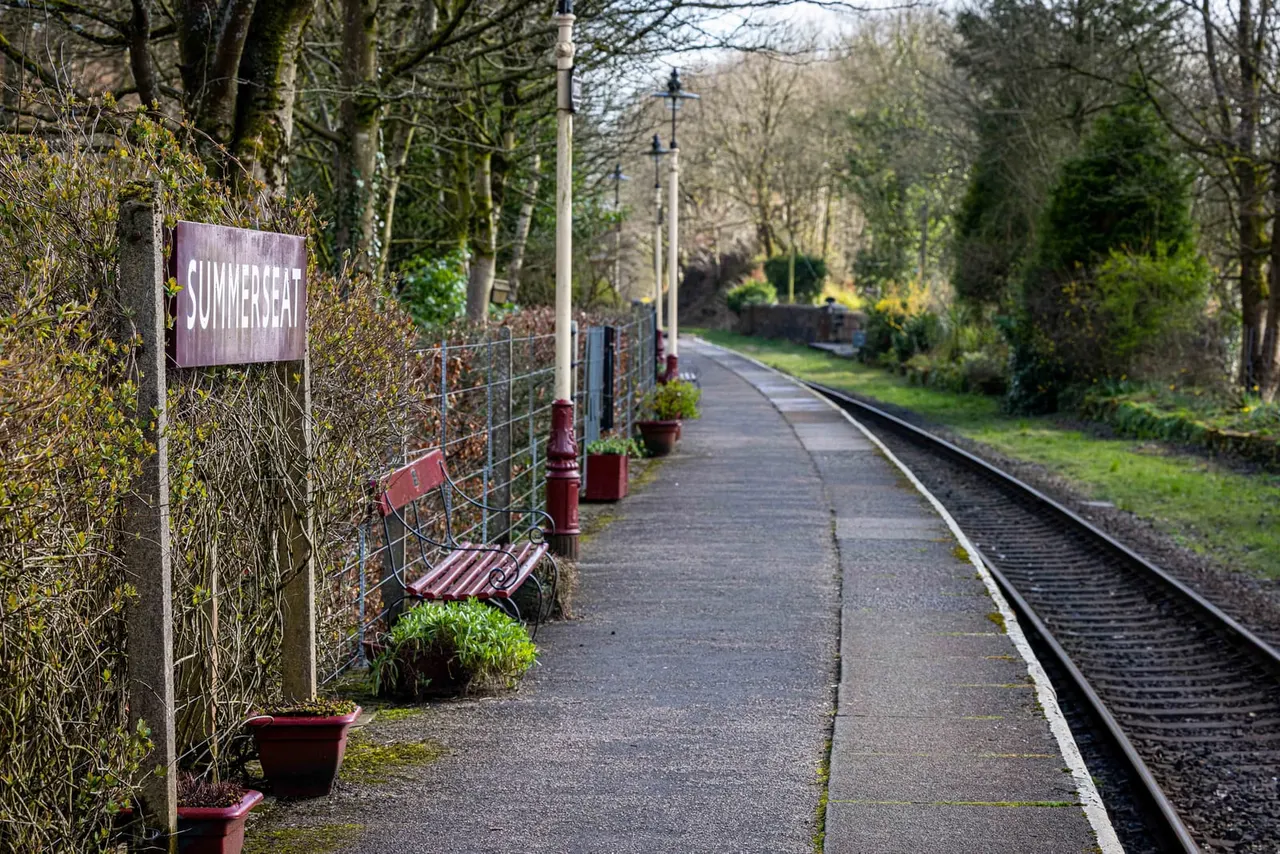 A railway station platform with benches, potted plants and a "Summerseat" sign, lined by bare trees and railway tracks.