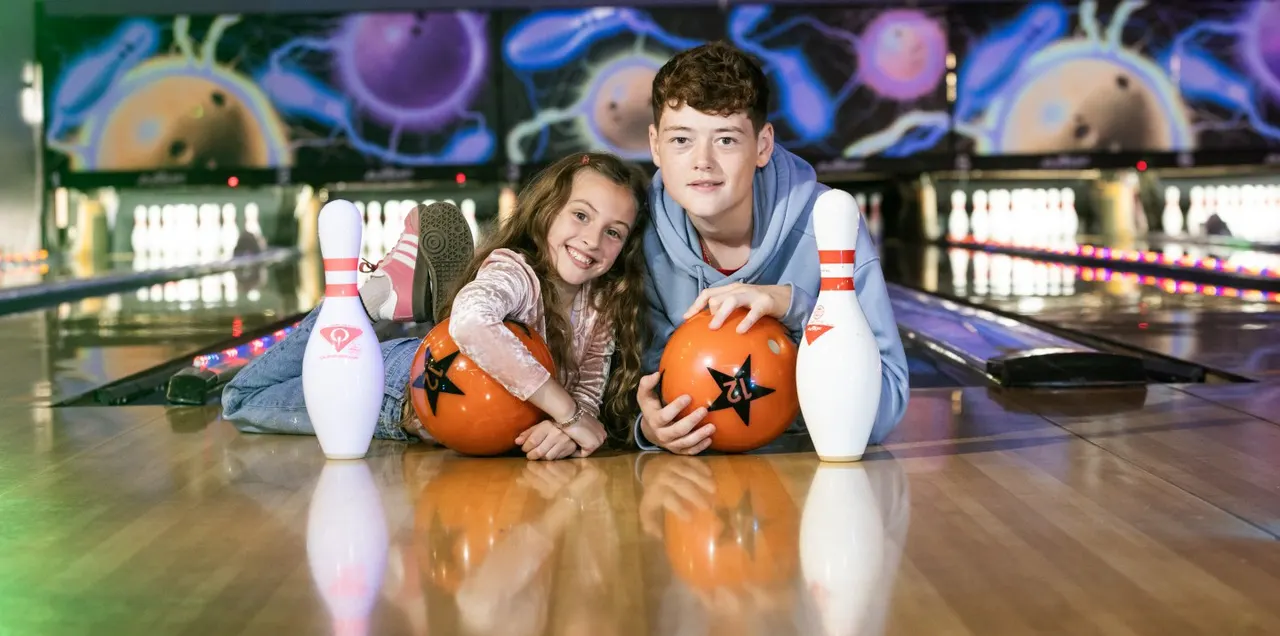 Two young people posing with orange bowling balls on a lane, surrounded by pins and colourful arcade graphics.