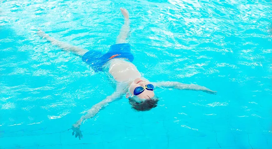 Child swimming butterfly stroke in bright turquoise pool wearing goggles and swim cap.