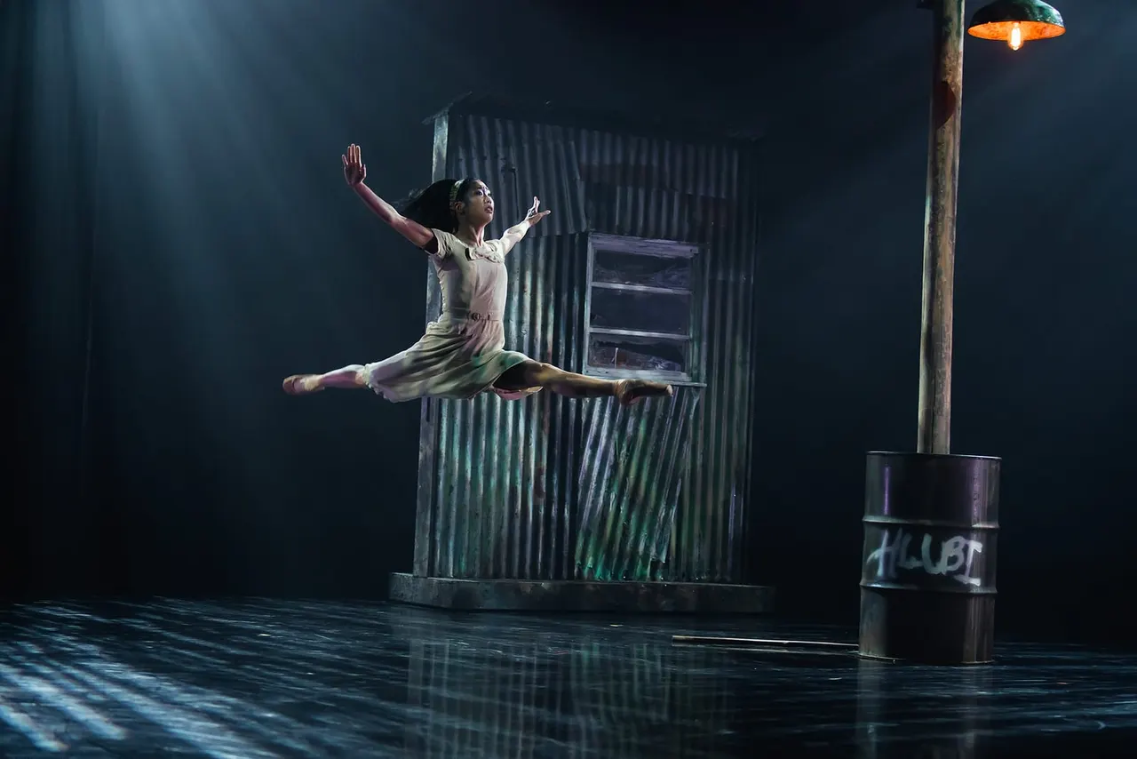 Dancer performing a leap in front of an industrial corrugated metal structure on a wet stage.