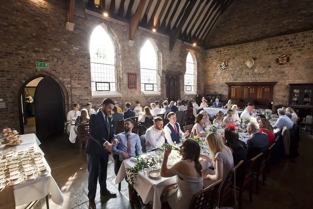 Wedding reception with guests seated at long tables in a stone-built historic hall with arched windows and wooden beams.