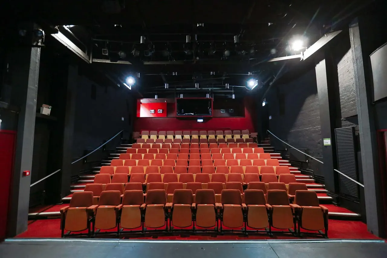 Empty cinema screen room with rows of red and orange seats facing a blank screen under bright lighting.