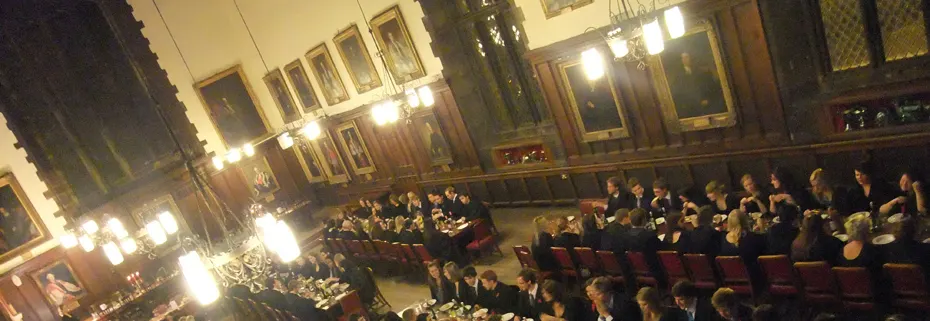 Historic wood-panelled hall with audience seated in red chairs facing a lit stage area during an evening event.