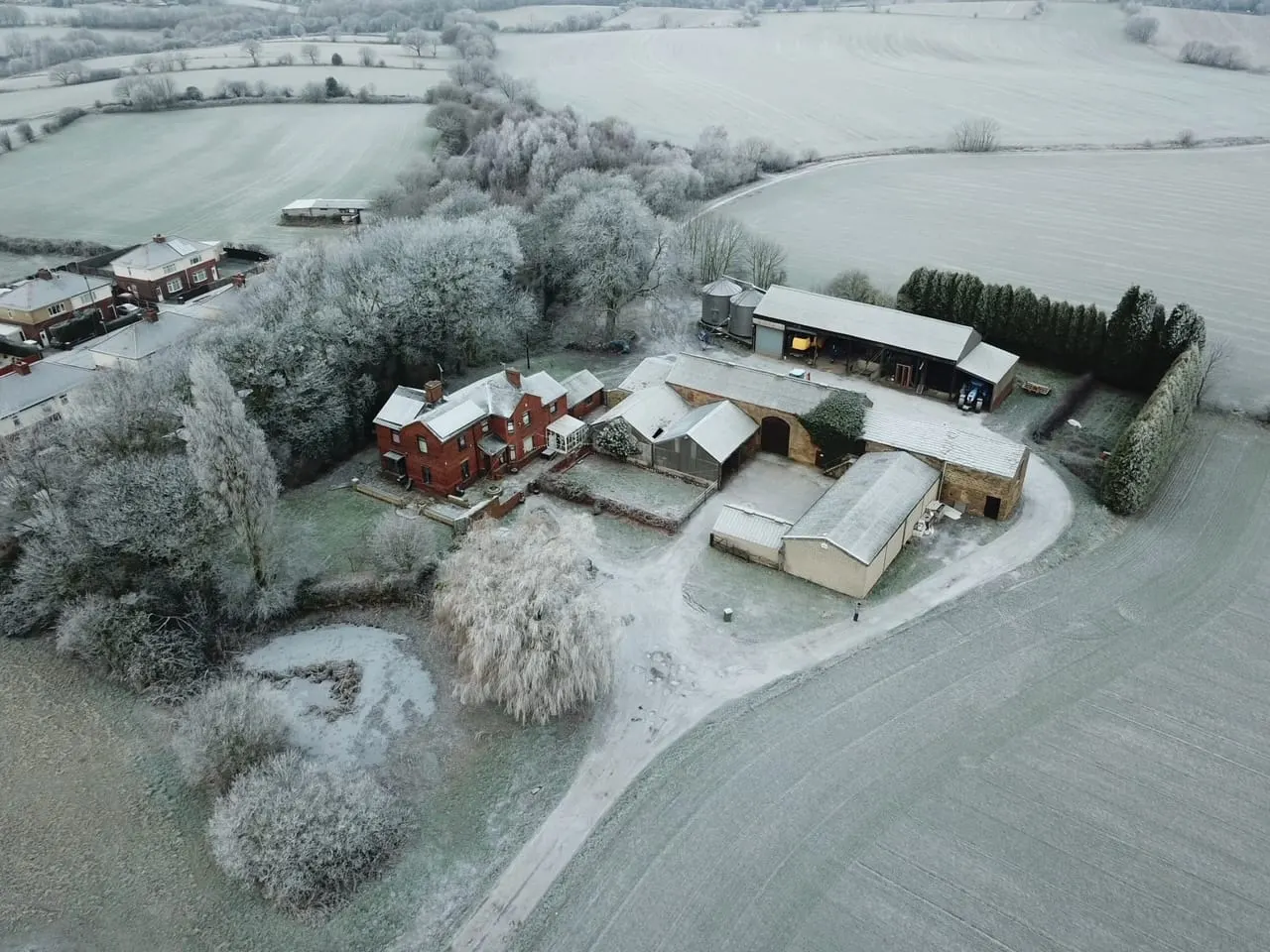 Aerial view of a frost-covered farm complex with a red brick house, barns and outbuildings surrounded by frozen fields and bare trees.