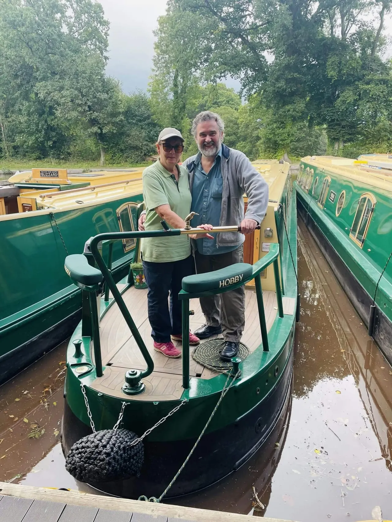Two people stand on the deck of a green narrowboat moored among other canal boats, surrounded by trees.