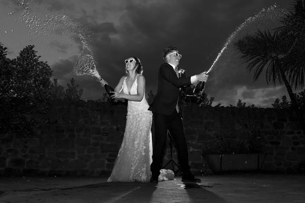 Bride and groom in formal wear spraying champagne with oversized props in a tropical garden setting.