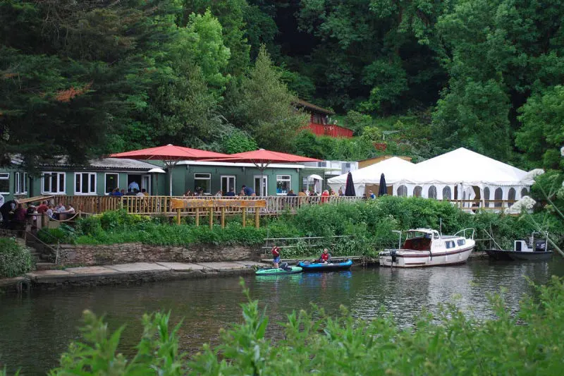 Riverside leisure venue with waterside buildings, red umbrellas, white marquee, moored boats and diners on a wooden terrace beside trees.