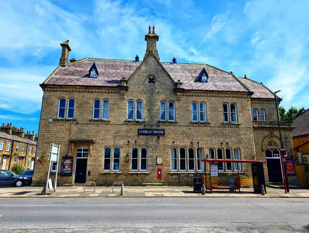 Victorian stone building housing Guiseley Theatre, with distinctive chimneys and blue-trimmed windows under a bright blue sky.