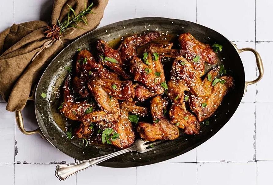 Glazed meat pieces with sesame seeds and herbs in a wok on a white tiled surface.