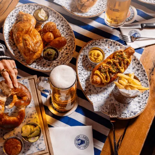 Overhead view of two plates of food with fried chicken, hot dogs with mustard, chips and accompaniments, alongside beers and condiments on a blue and white striped table.