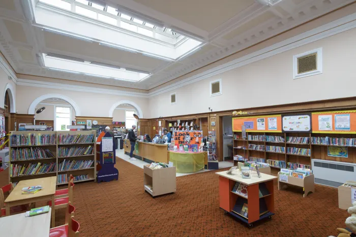 A bright, spacious children's library interior with bookshelves, display boards, skylights and colourful storage units, with a few visitors browsing.