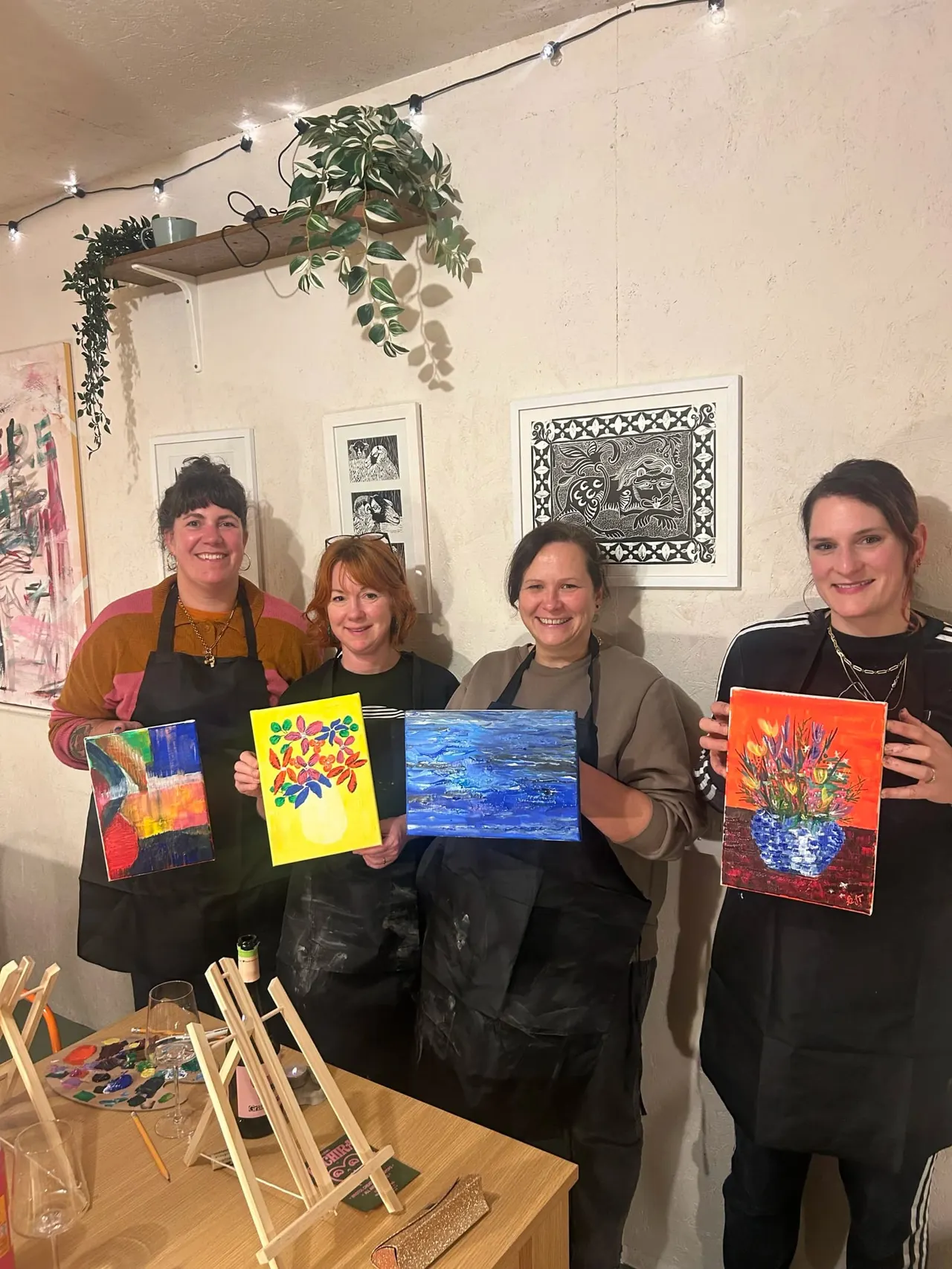 Four people in black aprons display their finished paintings at an art workshop, with easels and supplies on the table below.