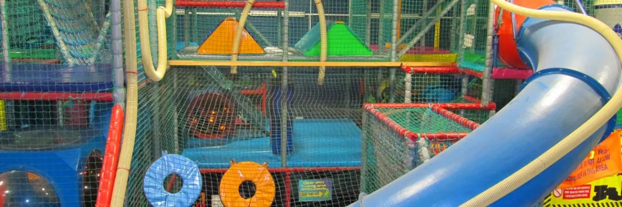 Colourful indoor soft play structure with slides, tunnels, netting and climbing equipment in bright primary colours.