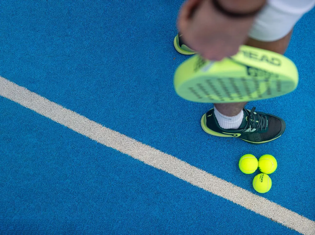 Person wearing neon trainers standing on a blue tennis court next to three yellow tennis balls and a white line.