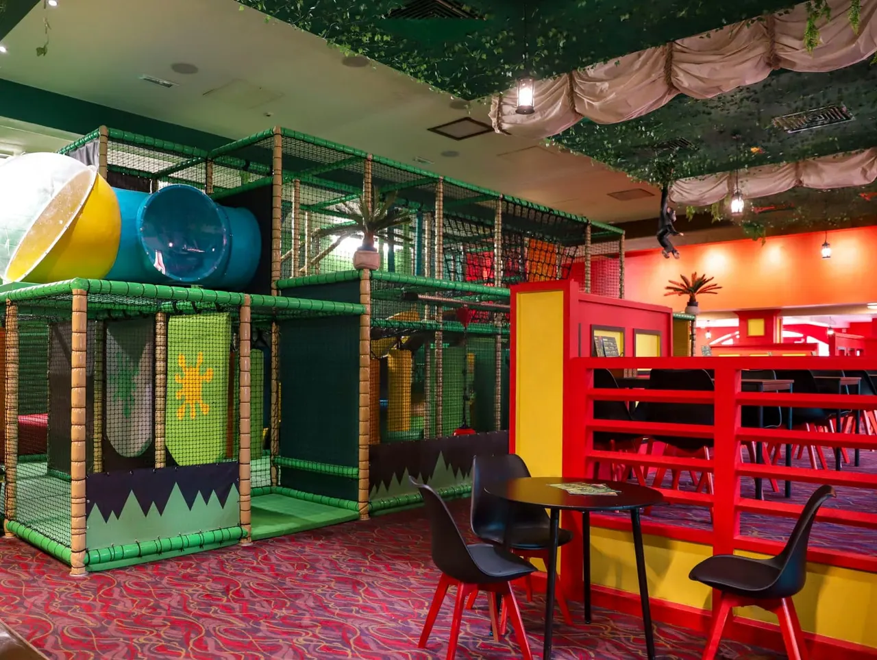 Indoor soft play area with green mesh climbing structure, slide and tunnels beside red shelving and seating area.