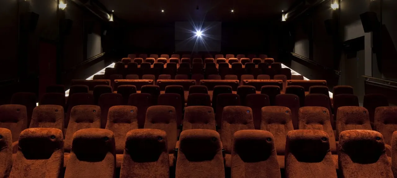 Empty cinema auditorium with rows of orange-brown seats facing a screen showing bright light.
