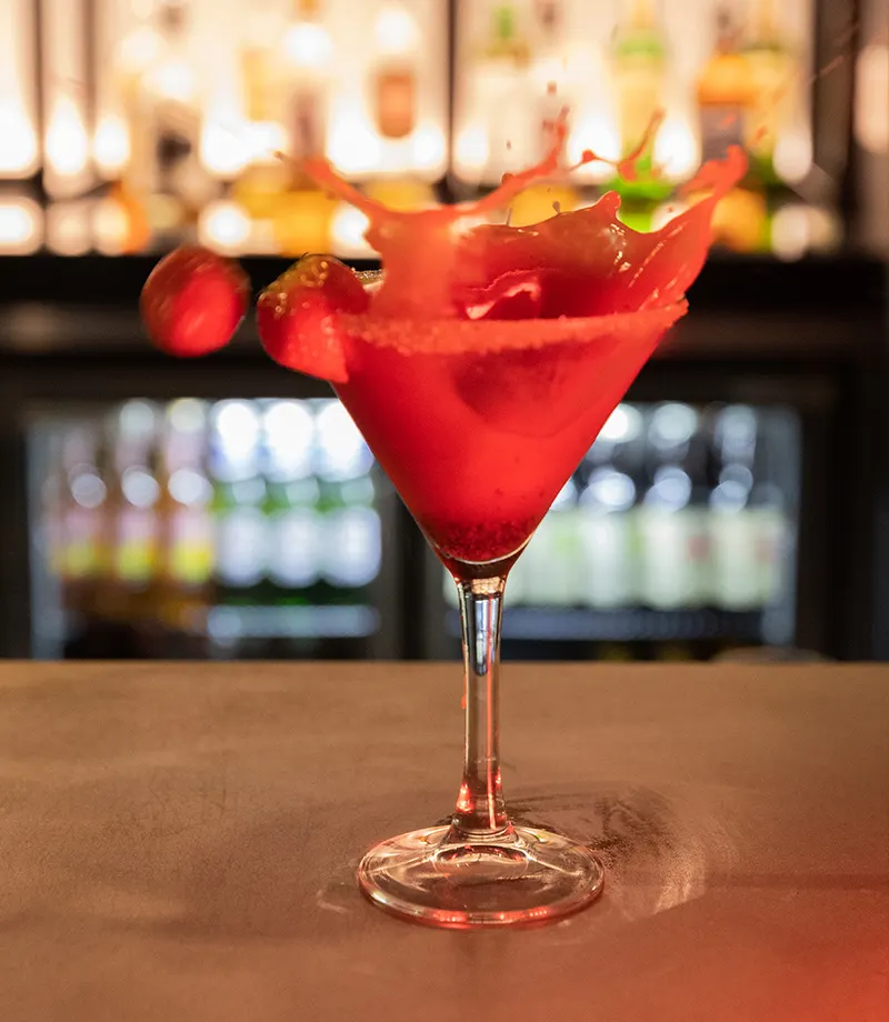 A red cocktail with splashing liquid and red pepper garnish sits on a bar counter with bottle-lined shelves blurred behind.