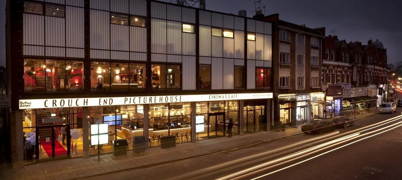 Illuminated cinema and café building at dusk with large windows, street frontage, and adjacent Victorian terraced houses.