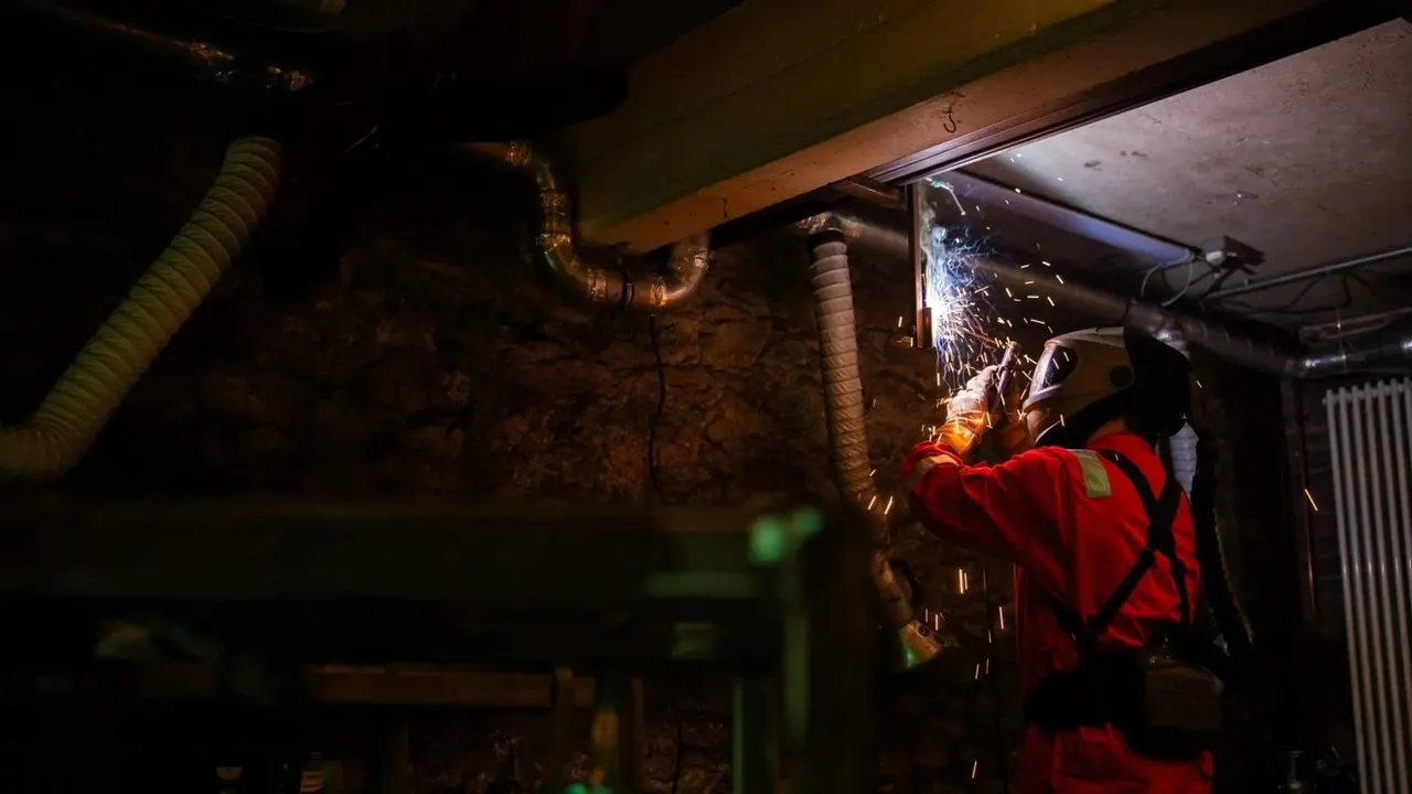 Worker in red protective clothing welding metal pipes in an industrial basement space.