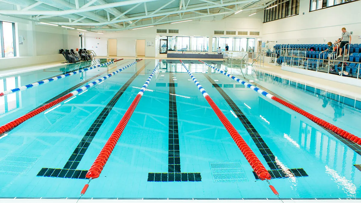 Indoor swimming pool with multiple lanes divided by red and white lane dividers, viewed from poolside with spectators in seating area.