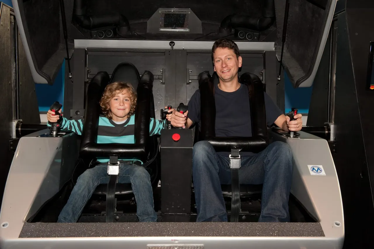 Adult and child on a motion simulator ride, holding control handles and smiling at the camera.