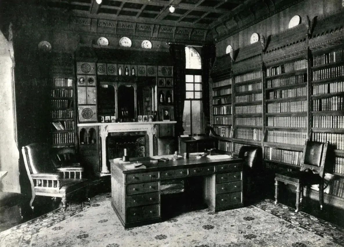 Historic library study room with floor-to-ceiling bookshelves, fireplace, desk and period furniture in black and white photograph.