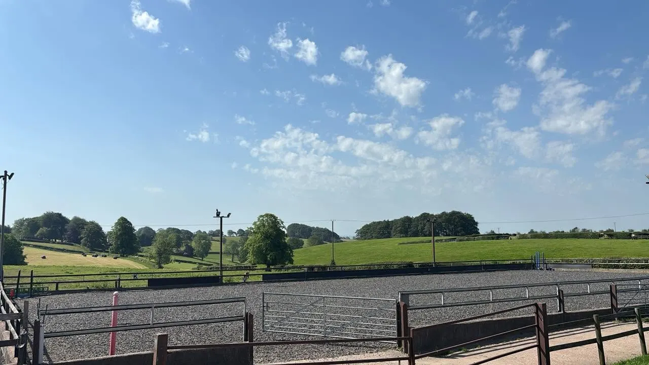 Empty equestrian arena with metal fencing and green countryside fields visible beyond under blue sky.