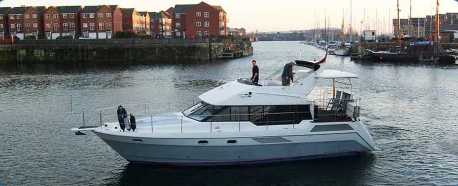 Two people on a motor yacht moored in a marina with historic waterfront buildings and sailing boats in the background.