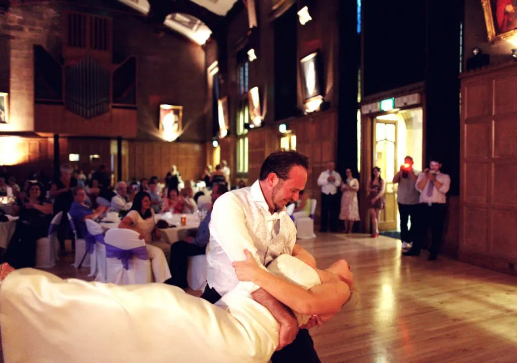 Man in white shirt dancing with a woman in a formal indoor venue with seated guests watching.