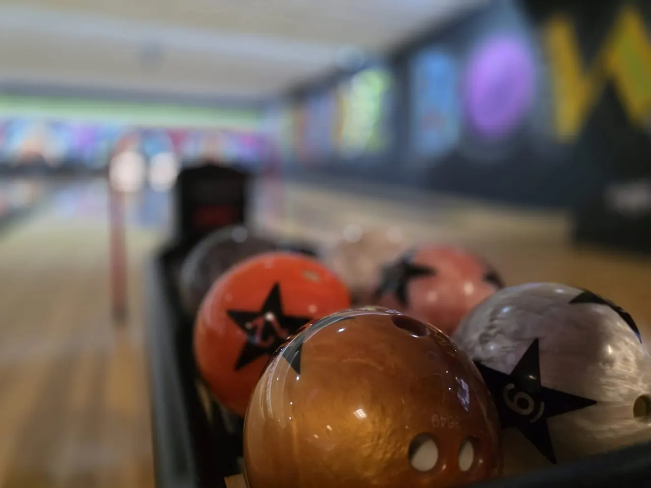 Bowling balls lined up on a lane at an indoor bowling alley with coloured pins visible in the distance.