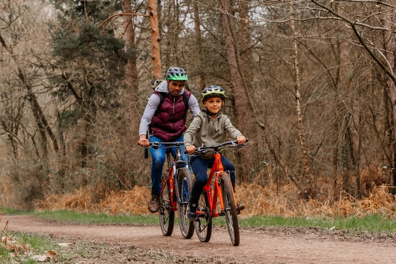 Adult and child cycling on a forest trail surrounded by bare trees and woodland.