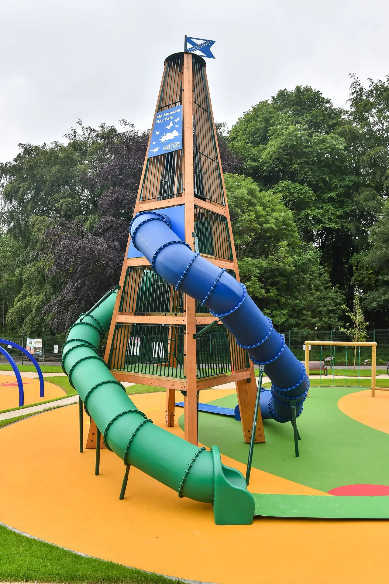 Tall wooden climbing tower with blue flag on top and green and blue tube slides spiralling down, surrounded by coloured rubber safety surfacing and trees.