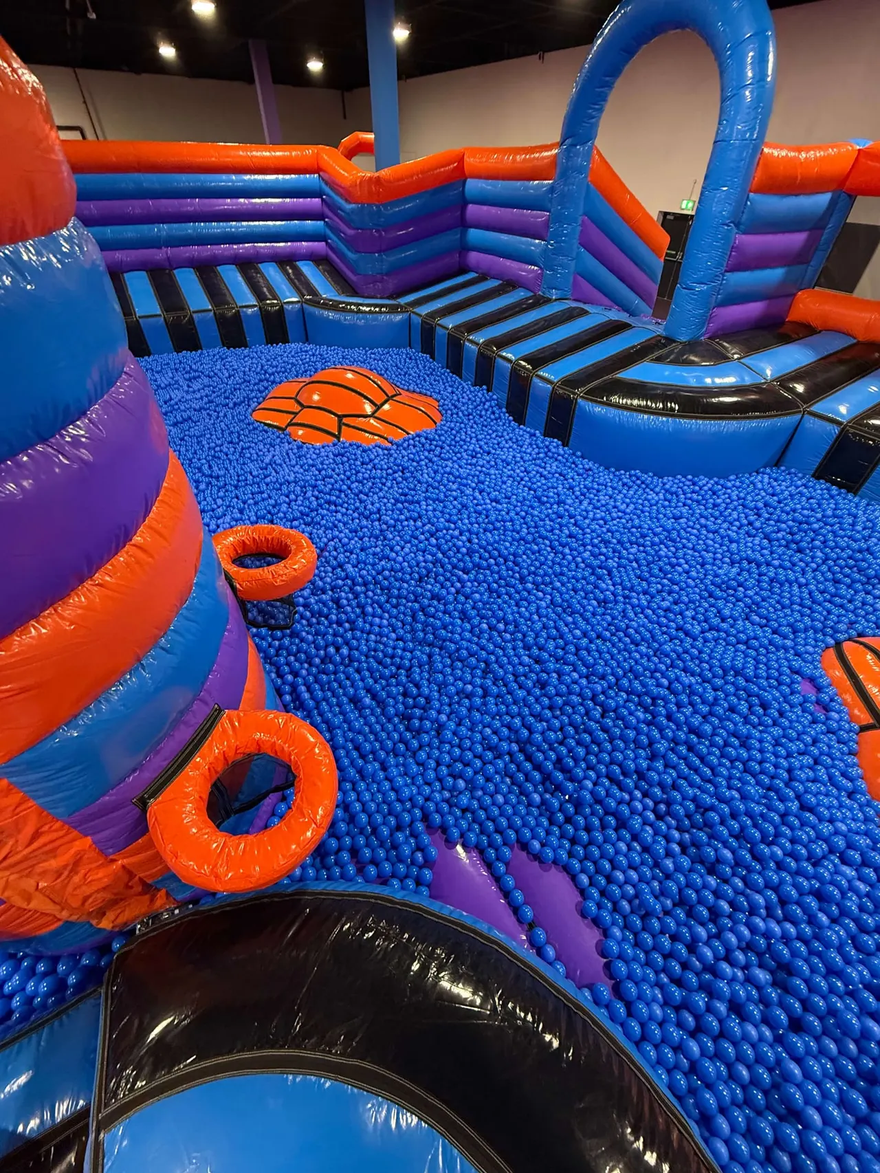 Inflatable play structure with blue ball pit, orange and purple padded walls, and basketball hoop design.