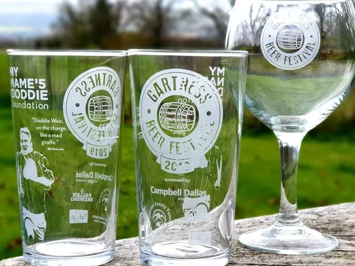 Three printed beer glasses and a wine glass displayed outdoors on a wooden surface with green fields behind.
