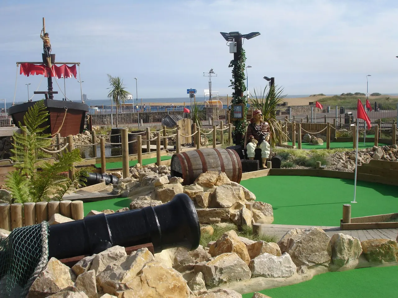Pirate-themed mini golf course with green fairways, rock features, cannons, and a ship structure overlooking a beach.