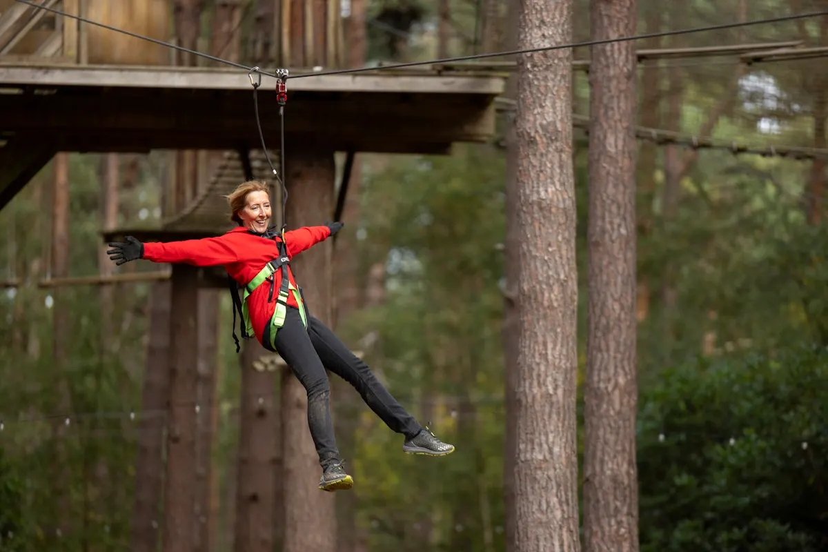 Woman zip-lining through a forest canopy course between tall trees, arms outstretched, wearing safety harness and red jacket.