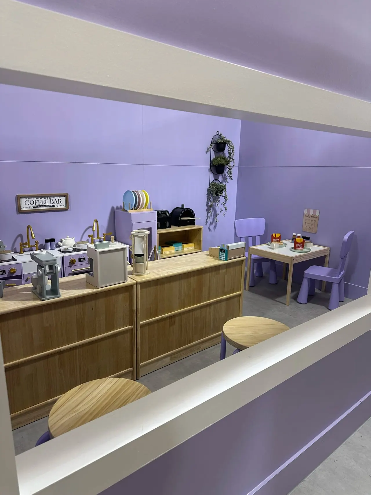 A play kitchen and café role-play space with purple walls, wooden counters, miniature appliances, seating areas, and decorative plants.