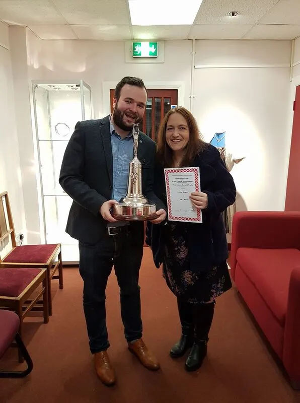 A man and woman smile at the camera whilst holding a trophy and certificate indoors.