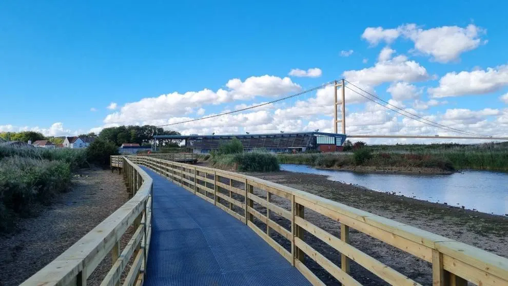 A wooden boardwalk with blue decking curves alongside a tidal estuary, with a suspension bridge and industrial buildings visible across the water.