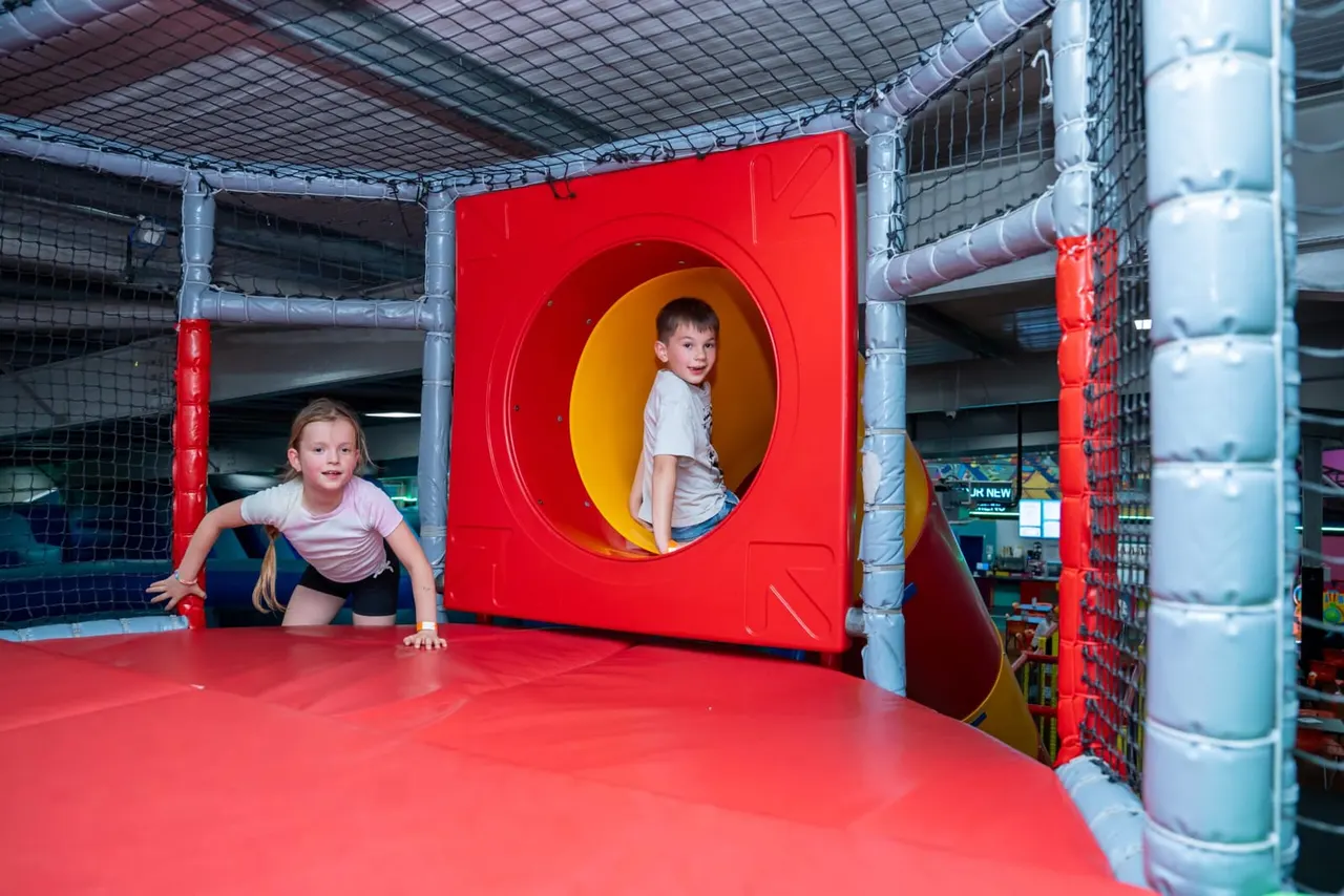 Two young children playing in a soft play structure with red panels and yellow tunnels indoors.