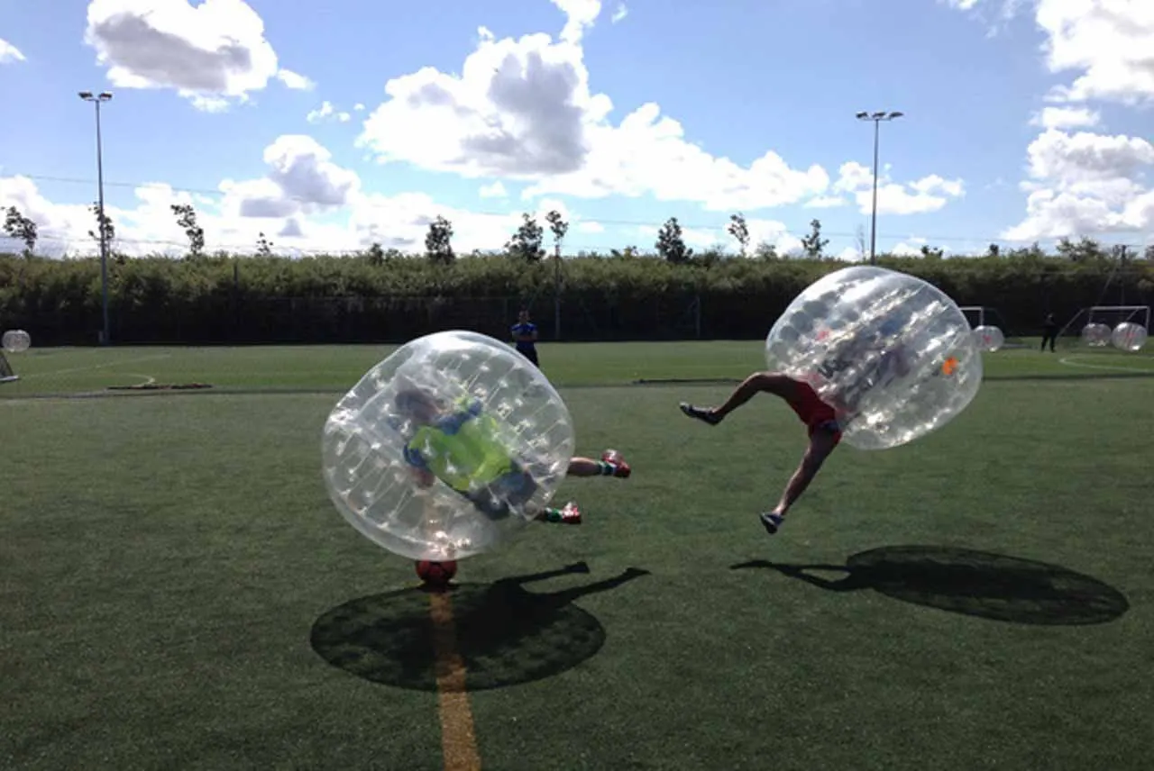 Two people inside large inflatable bubbles playing bubble football on a grass pitch.