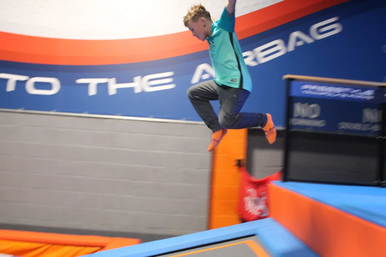 Child jumping on a colourful trampoline with padded obstacles inside an indoor trampoline park.